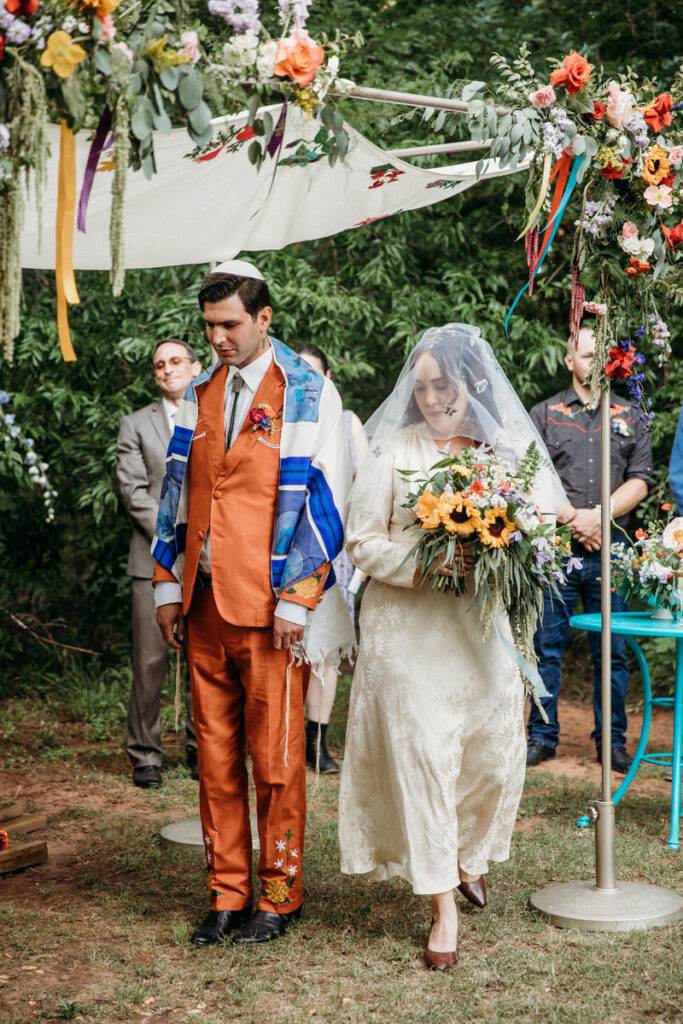Groom in orange suit and bride in vintage dress standing under fabric canopy with wedding guests in background