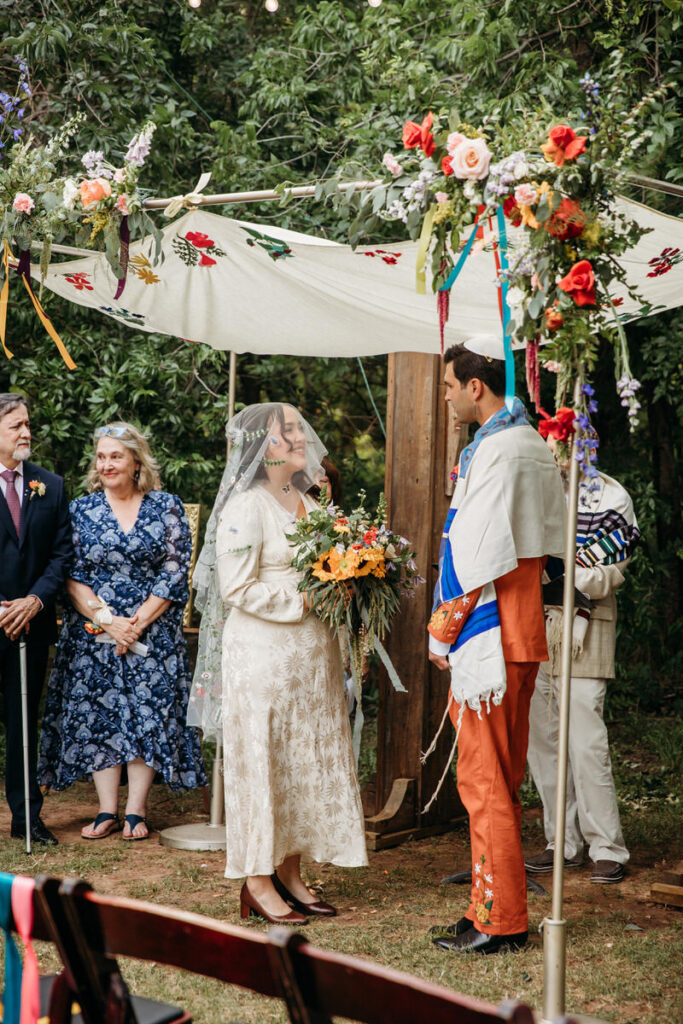 Groom in orange suit and bride in vintage dress smiling at each other under fabric canopy with wedding guests behind them