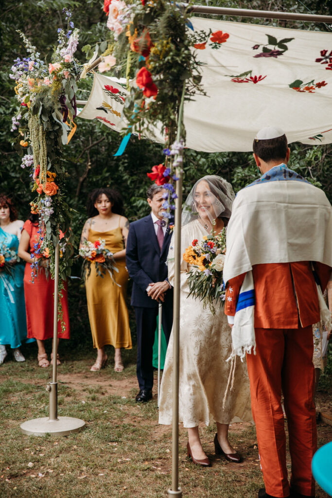 Bride in vintage dress and groom in orange suit under fabric canopy with bridesmaids in multicolor dresses in background