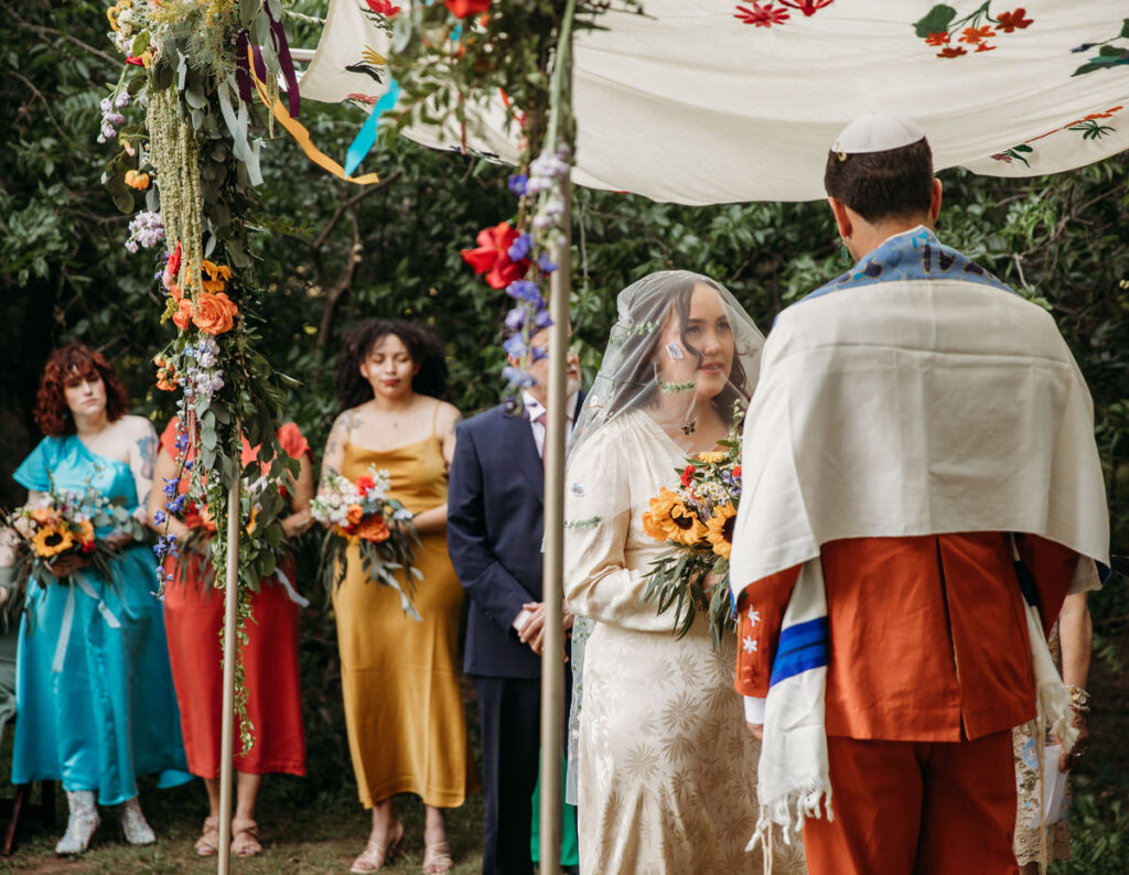 Groom in orange suit and bride in vintage dress during wedding ceremony with bridesmaids in background