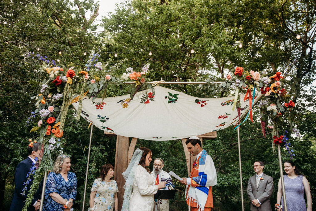 Groom in orange suit and bride in vintage dress standing under fabric canopy reading their vows