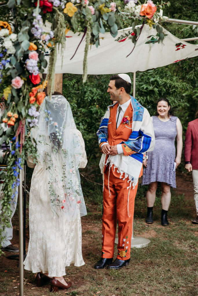 Groom in orange suit and bride in vintage dress standing under fabric canopy looking at rabbi