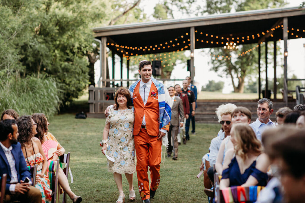 A groom in an orange suit with a blue floral shawl walks arm-in-arm with a woman in a floral dress at a wedding ceremony, with guests seated in a green outdoor setting and string lights above