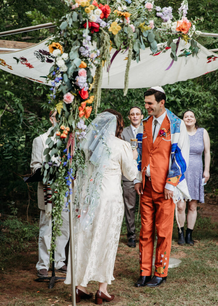 Groom in orange suit and bride in vintage dress standing under fabric canopy adorned with flowers
