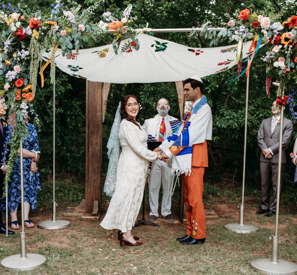 Groom in orange suit and bride in vintage dress stand under fabric canopy while bride looks out to the guests
