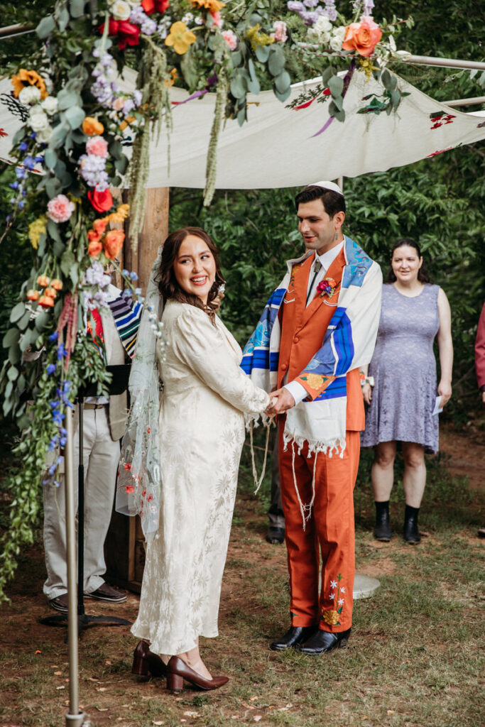 Groom in orange suit and bride in vintage dress standing under fabric canopy with bride looking behind her