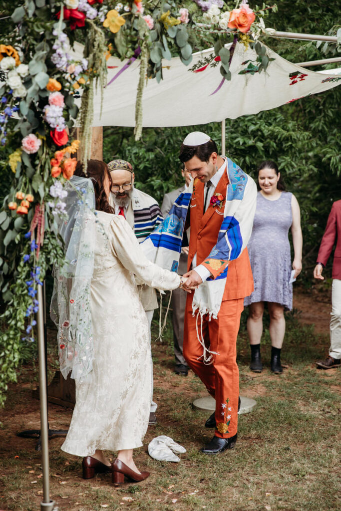 Groom in orange suit and bride in vintage dress standing under fabric canopy during wedding ceremony