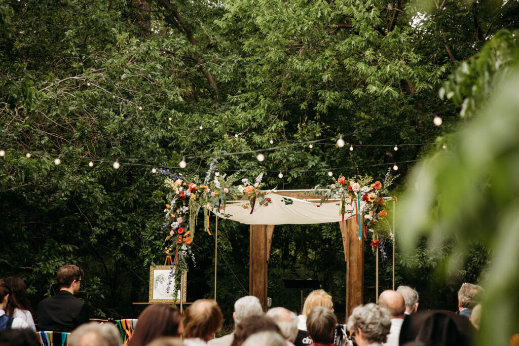 A floral-decorated wedding arch with a sheer fabric banner set up outdoors, with greenery in the background and guests seated in anticipation of the ceremony