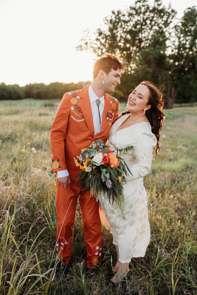 Groom in orange suit and bride in vintage dress standing in field looking at each other affectionately