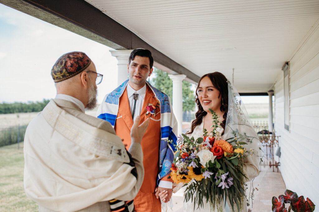 A bride and groom holding hands, facing a rabbi on a covered porch during their wedding ceremony, with the groom in a colorful tallit and the bride smiling, holding a vibrant bouquet