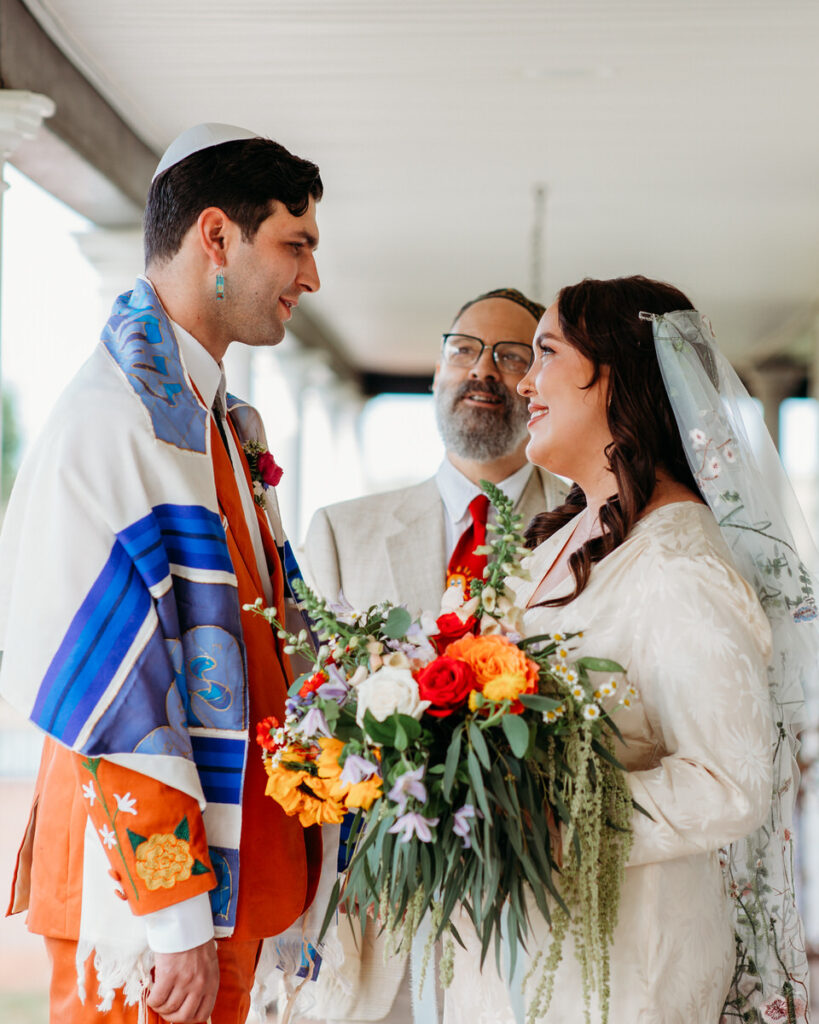 A bride and groom standing under a porch roof with an officiant, exchanging smiles and vows during an intimate wedding ceremony