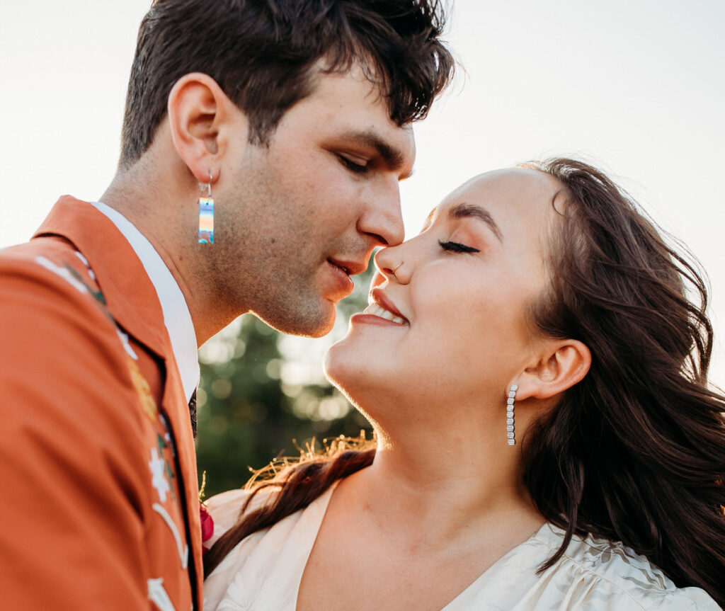 Close-up of a bride and groom about to kiss, with the groom in a bright orange suit and the bride in a white dress, their faces partially illuminated by the golden hour sunlight