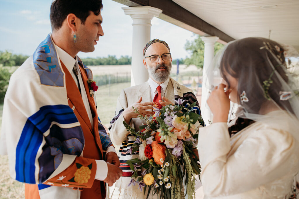 A bride and groom standing under a porch roof with an officiant, exchanging smiles and vows during an intimate wedding ceremony