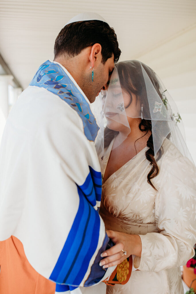 A tender moment as a groom in a white yarmulke and tallit leans toward his veiled bride, both in profile