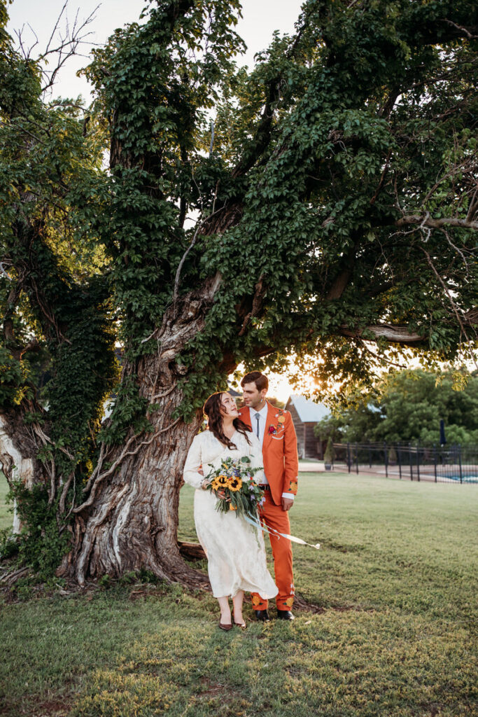 Groom in orange suit and bride in vintage dress standing under intertwined tree looking at each other affectionately