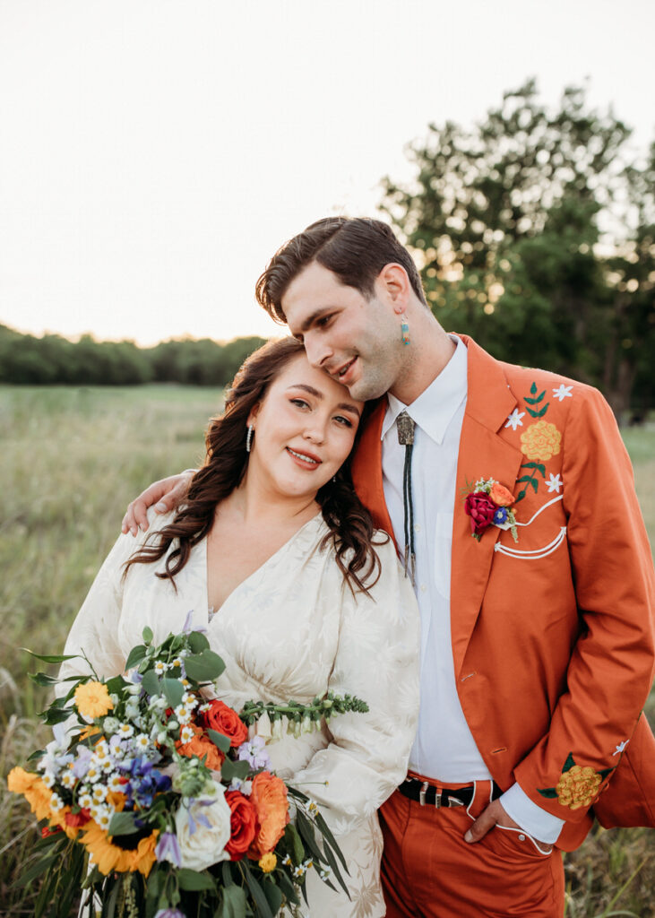 A groom in an ornate orange suit and a bride in a floral embroidered gown share a serene moment in a grassy field, with the sunset casting a warm glow on their faces