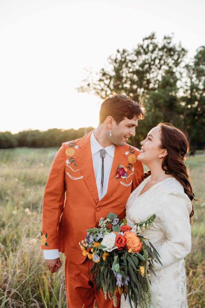 Groom in orange suit and bride in vintage dress standing in field embracing and looking at each other affectionately