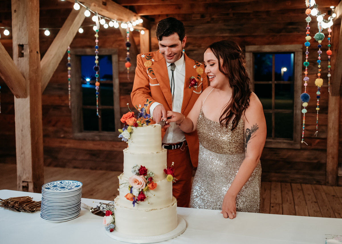 Groom in orange suit and bride in sparkly dress cut wedding cake adorned with flowers