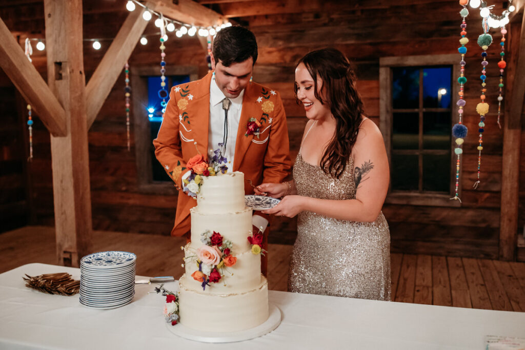 Groom in orange suit and bride in sparkly dress cutting wedding cake adorned with flowers