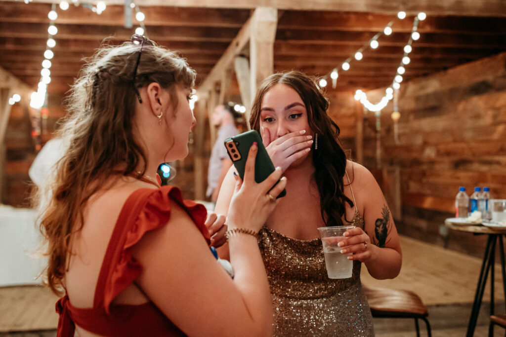 A surprised bride in a glittering gold dress covers her mouth with her hand while holding a drink as she looks at a smartphone screen shown by a guest at a wedding reception