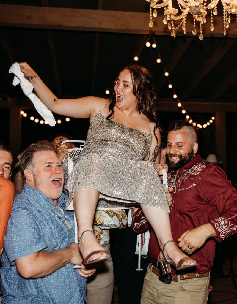 Bride in sparkly dress being lifted in chair by two men