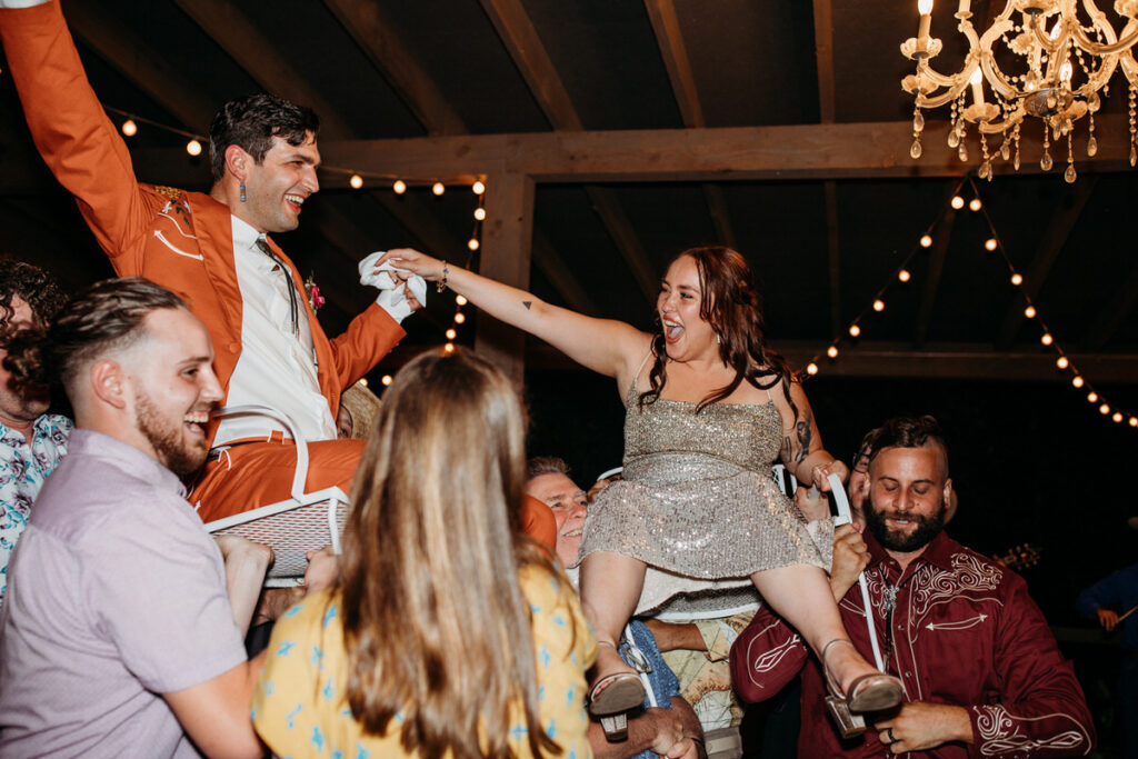 Groom in orange suit and bride in sparkly dress being lifted in chairs by weddings guests