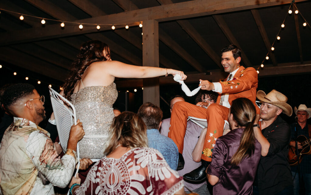 A bride in a sparkling gold dress reaching out to a groom in an orange embroidered suit as they are hoisted on chairs by wedding guests, with string lights above at an evening reception