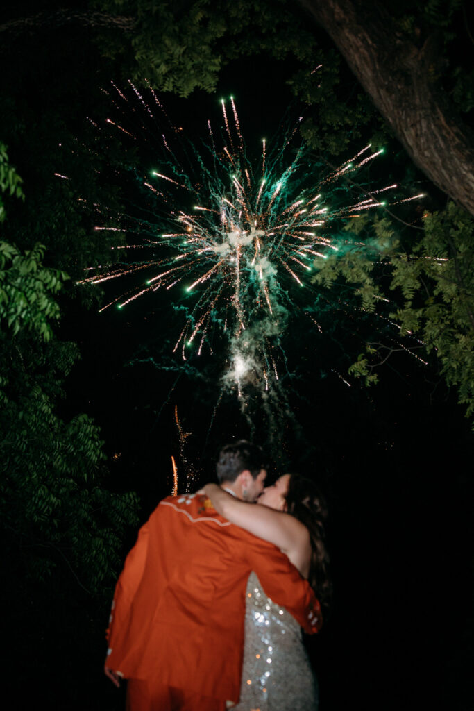 Groom in orange suit and bride in sparkly dress embrace and kiss under fireworks