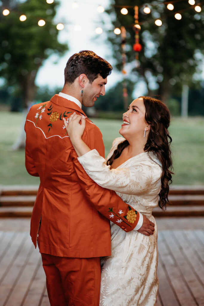 Groom in orange suit and bride in vintage dress dancing on wood floor under string lights