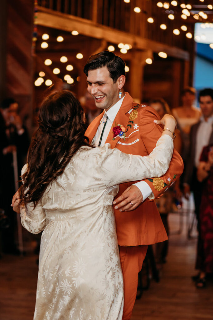 Groom in orange suit and bride in vintage dress dress under string lights in rustic barn