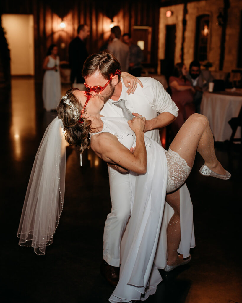 A joyful bride in sunglasses dips dramatically in a groom's arms on the dance floor, showcasing a lively and spontaneous moment at their wedding reception