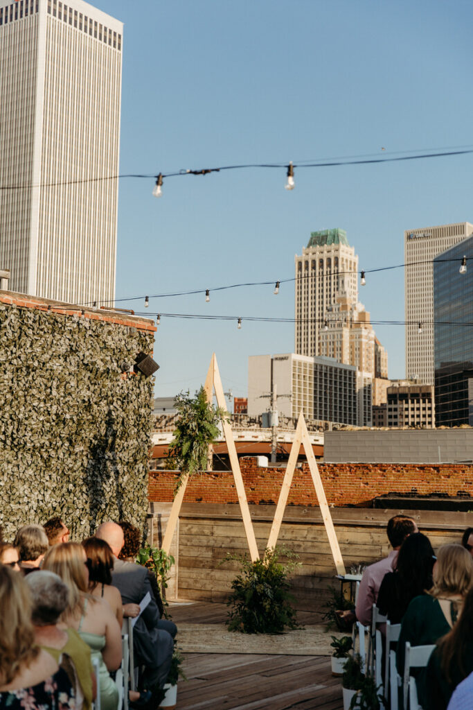 Guests seated at a rooftop wedding ceremony with urban skyline in the background, featuring string lights and a wooden triangular arch