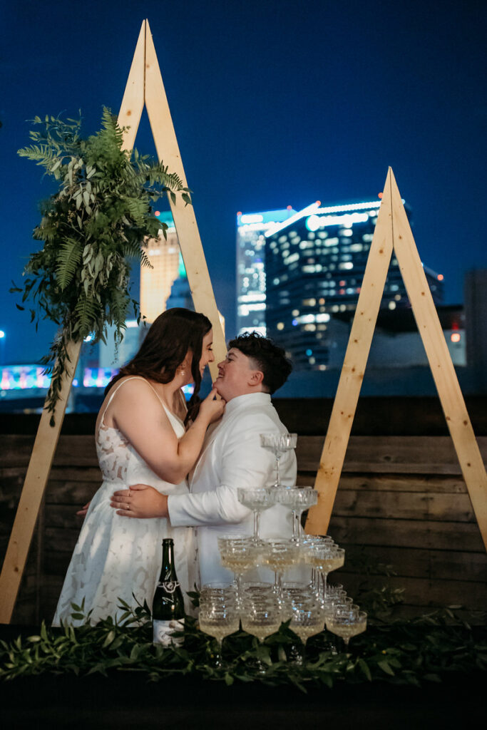 Bride and partner sharing an intimate moment under a triangular wedding arch on a rooftop, with city lights behind them