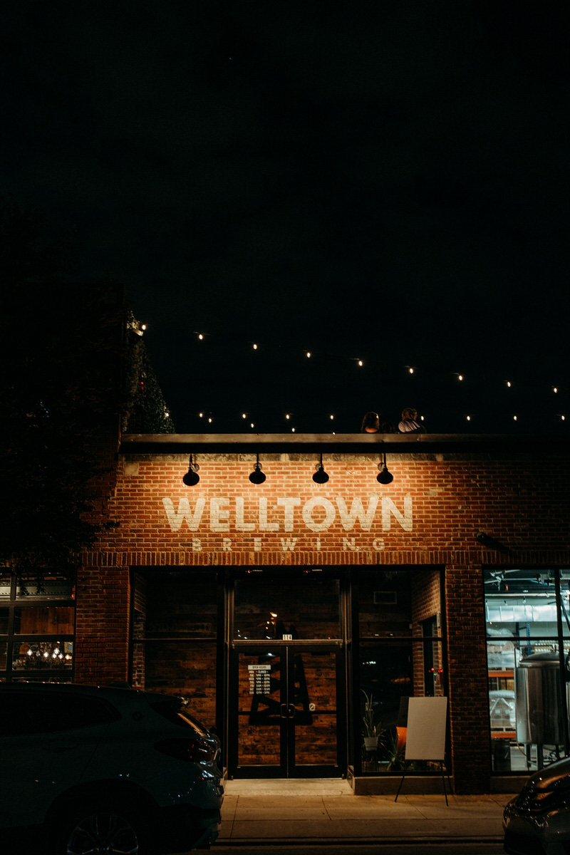 Exterior night view of Welltown Brewing with its name in large letters on the brick façade, lit by overhead string lights and street lamps