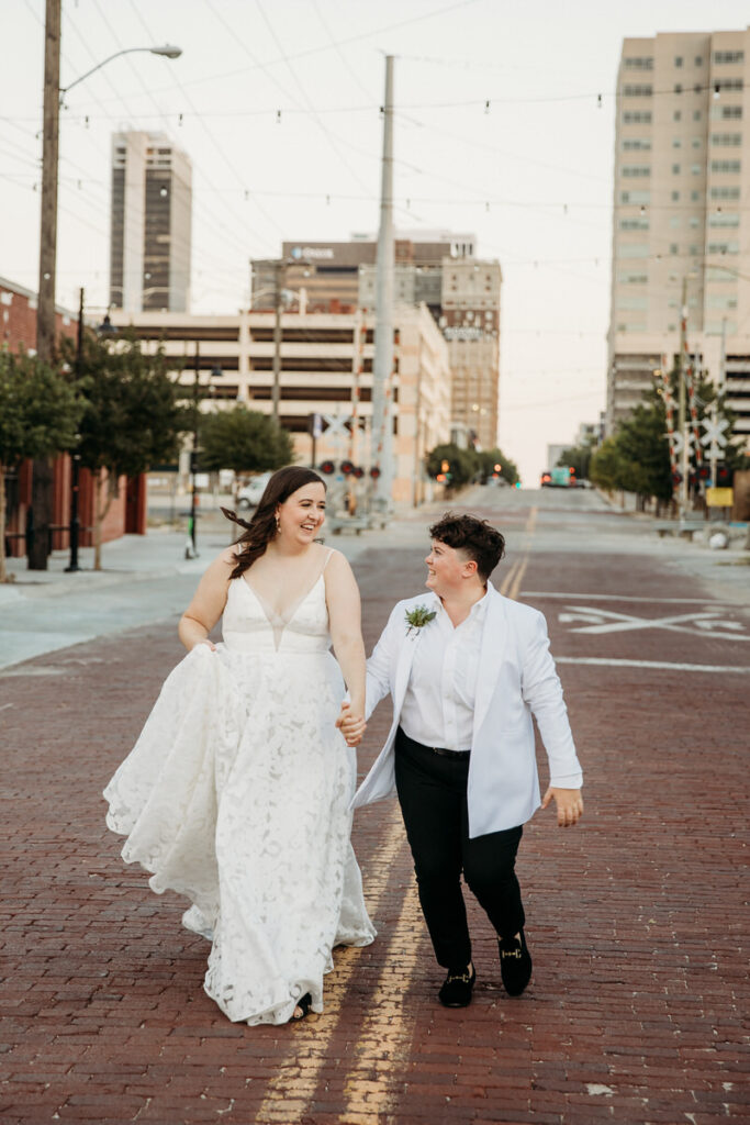 Bride in a flowing white dress and partner in a white jacket and black trousers walking hand in hand on a city street, smiling at each other, with urban buildings and string lights in the background