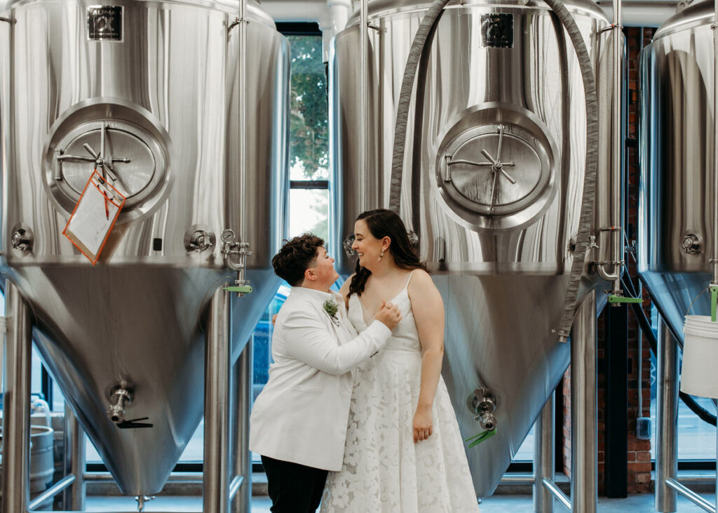 A couple in wedding attire standing in front of stainless steel brewing tanks, looking into each other's eyes with affection