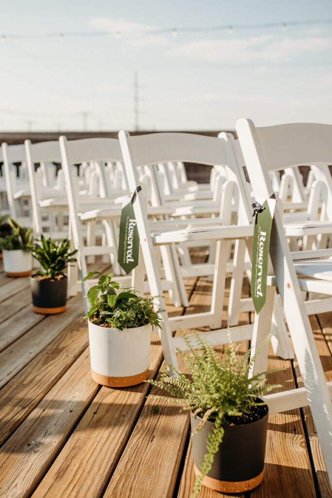 Reserved seating at a rooftop wedding venue with white chairs and potted green plants on a wooden deck, under a clear blue sky