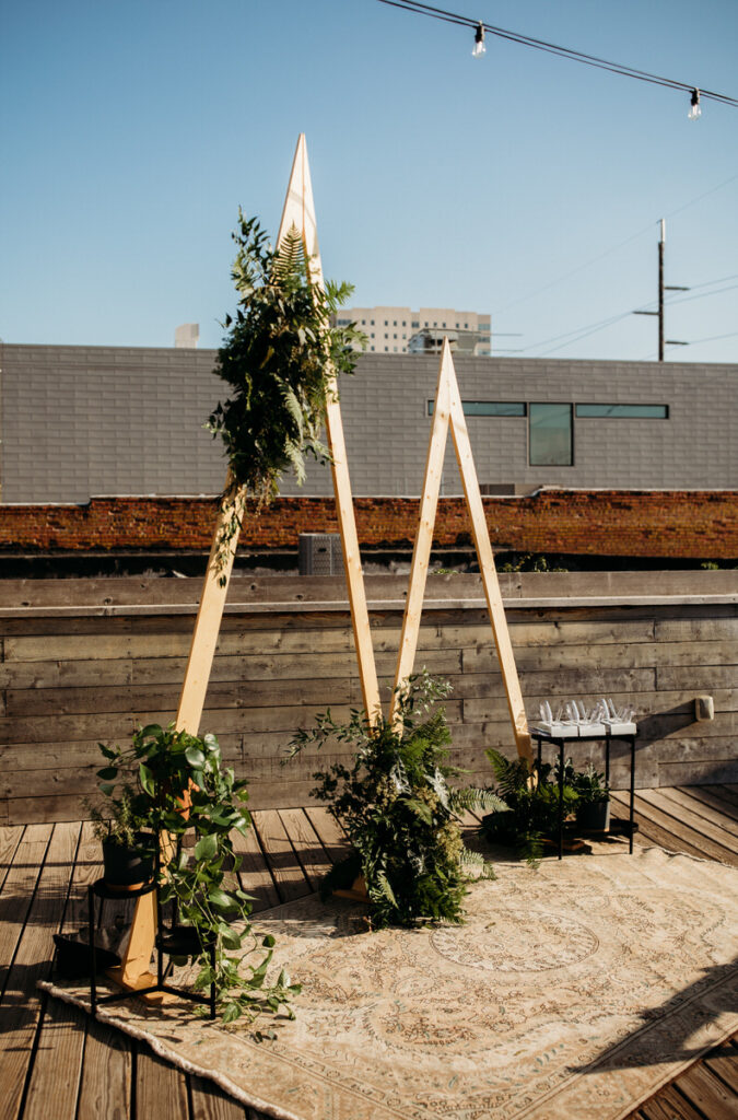 A simple yet elegant outdoor wedding setup featuring a wooden arch adorned with greenery and a vintage-style rug, with the cityscape in the distance