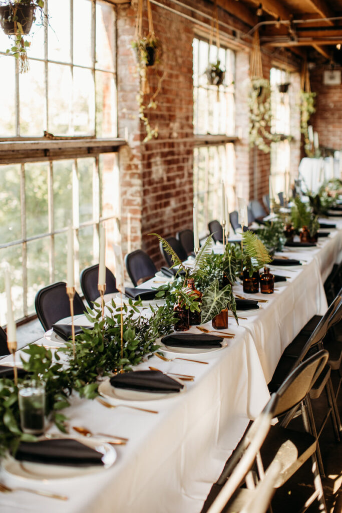 Long reception table elegantly set with black plates, gold cutlery, and a garland of greenery, next to large windows in a brick-walled venue