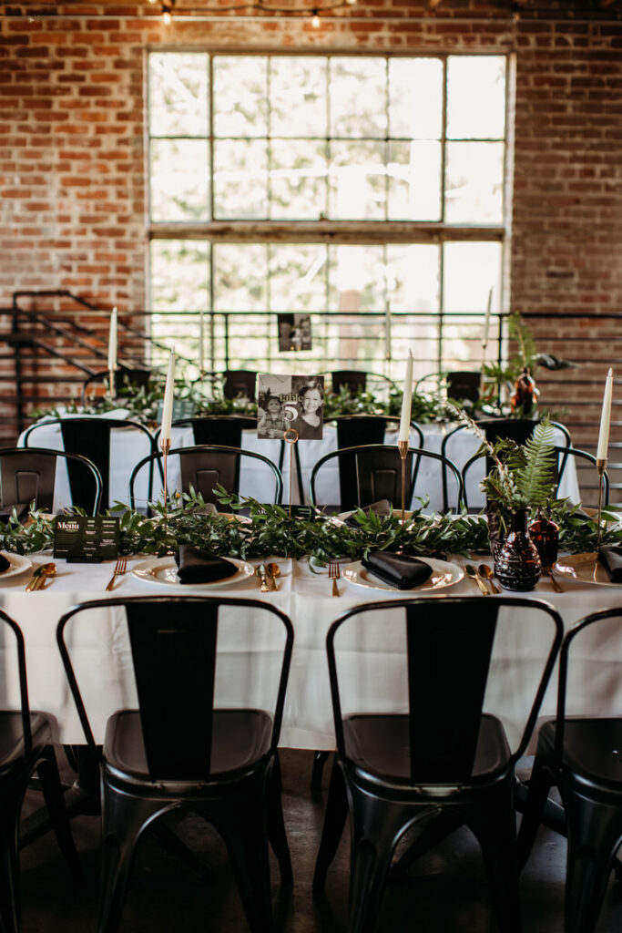 Elegant wedding table setting featuring black chairs, white tablecloth, and natural greenery centerpieces, with personalized table cards