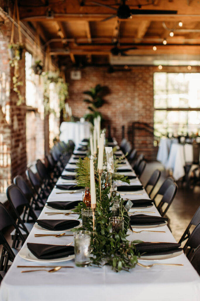 tylish wedding reception table with black and gold place settings and a lush greenery runner, in an industrial-chic space with exposed beams