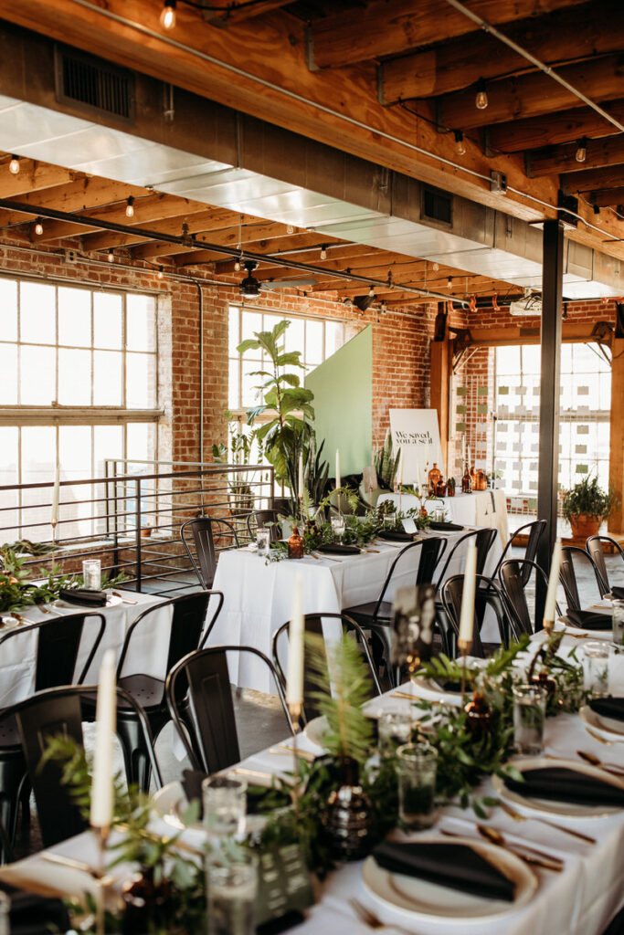 Wedding reception venue with tables set with white linens, green plants, and a large green wall accent, in a rustic space with wooden beams overhead