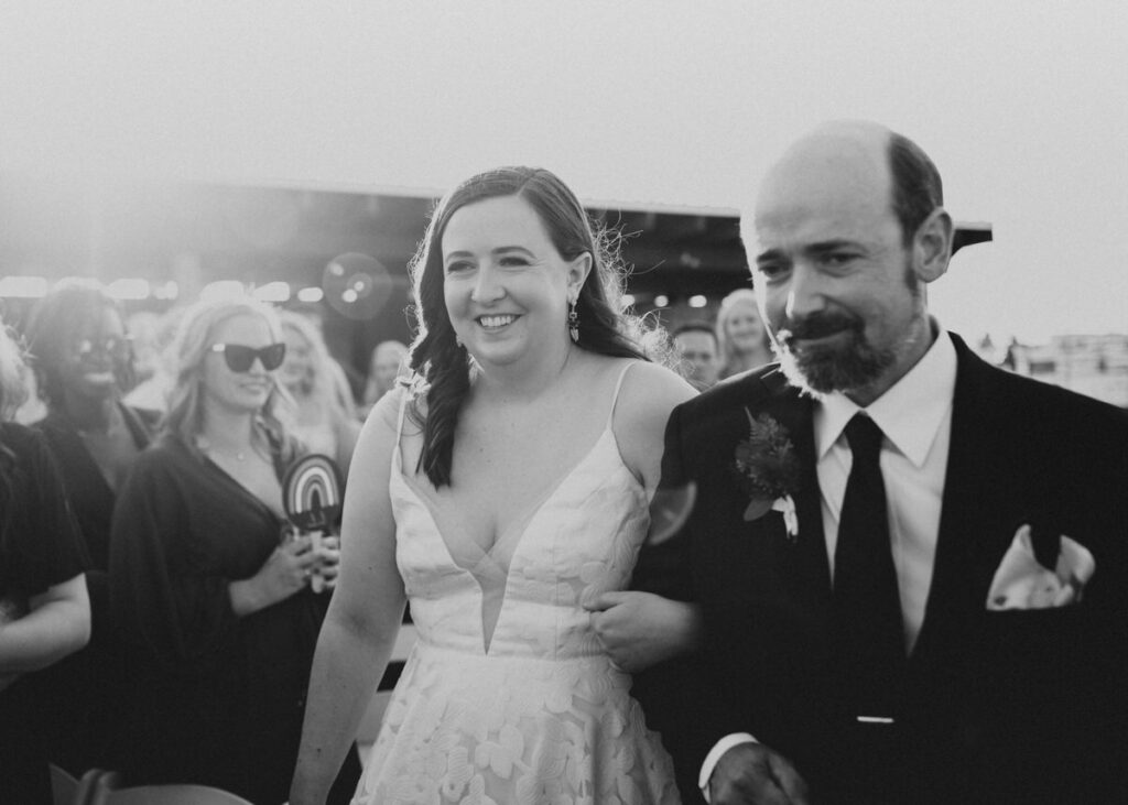 Black and white photo of a bride smiling broadly as she walks with an older man in a suit, both looking ahead, with guests in the background