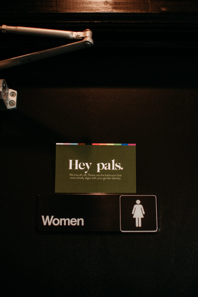 Inclusive bathroom sign reading 'Hey pals. We love all y'all. Please use the bathroom that most closely aligns with your gender identity,' mounted below a standard 'Women' restroom sign