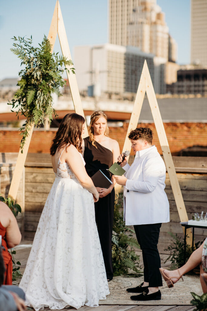 Bride in a white gown and partner in a white jacket and black pants standing during a rooftop wedding ceremony, with an officiant and guests in the background