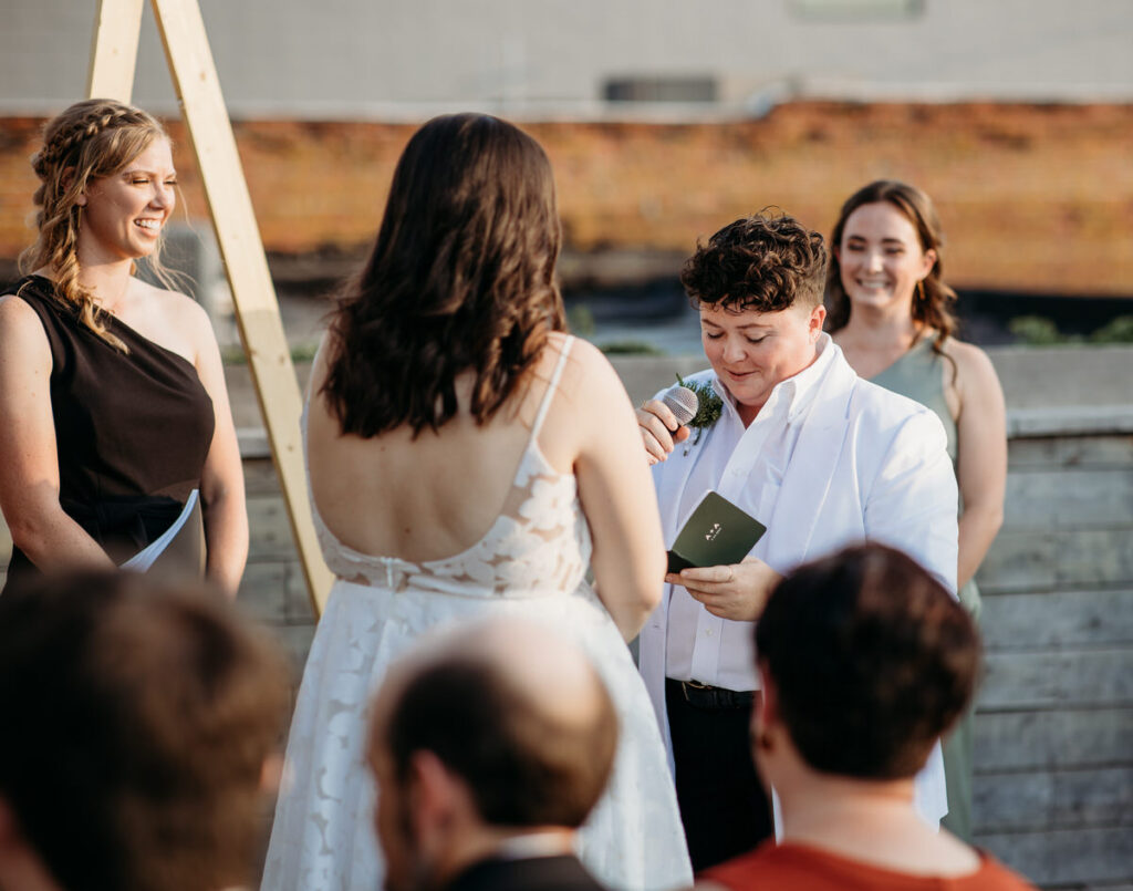 A wedding ceremony on a rooftop with a bride and partner facing an officiant, backdropped by a simple wooden arch and the urban skyline