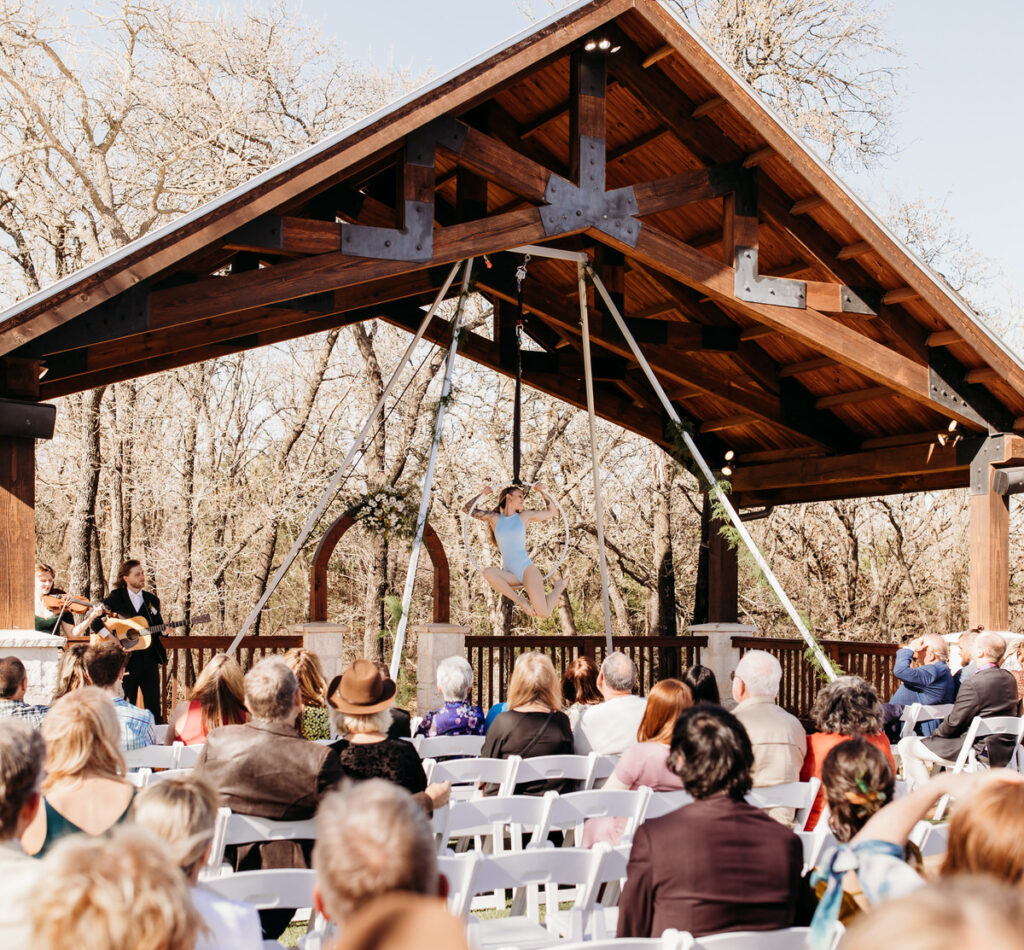 Aerial silks performance during an outdoor wedding ceremony, with guests seated and watching attentively under a rustic wooden pavilion