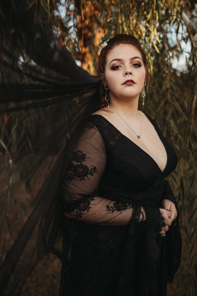 A woman in a black lace dress with sheer sleeves stands confidently among weeping willow branches, embodying a moody elegance
