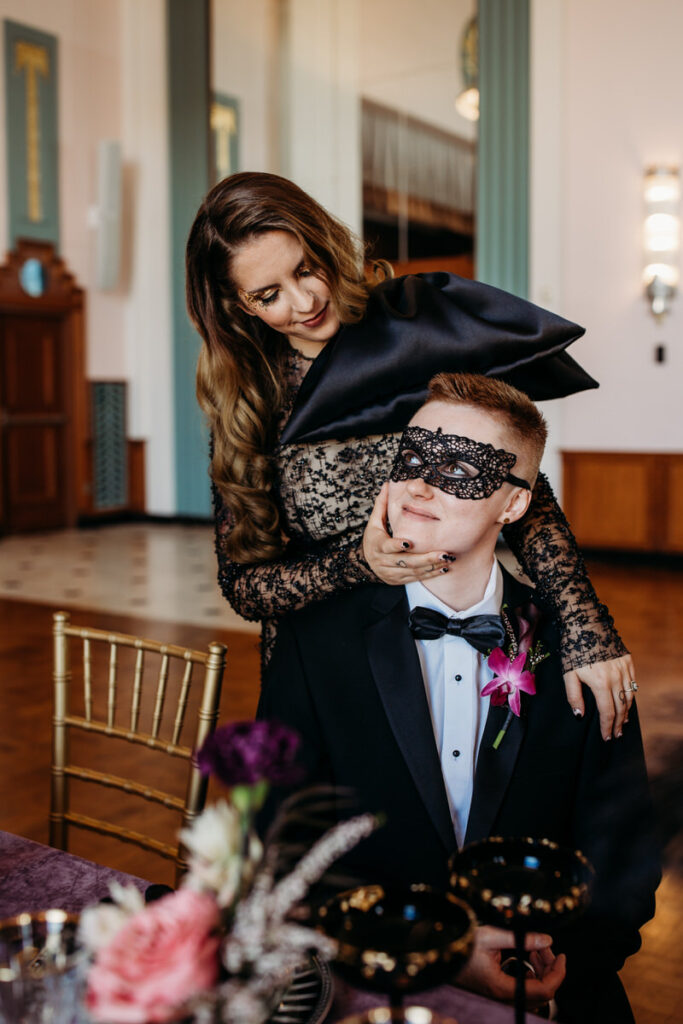 A couple in a playful masquerade moment, the woman in a lace black dress, gently lifts a mask from the man's eyes, revealing a tender gaze