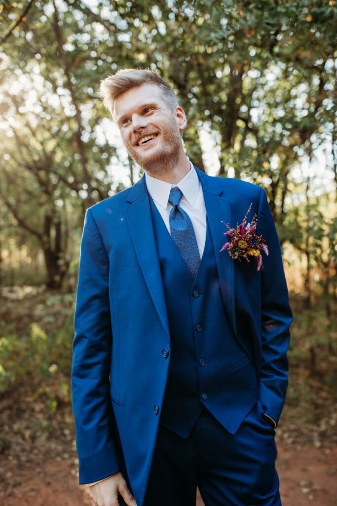 A groom in a vibrant blue suit smiles broadly, set against a lush forest backdrop, his boutonniere adding a pop of pink and green